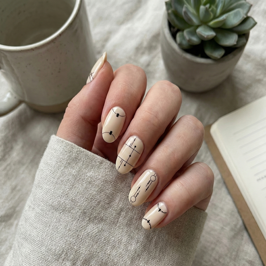 Hand with beige nail polish and black arrow design, cup, and plant in the background