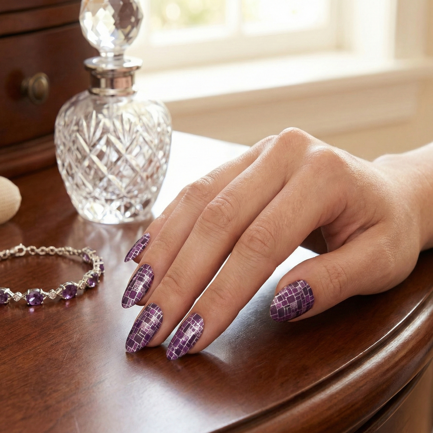 Hand with purple checkered nail design on a wooden surface with a crystal perfume bottle.